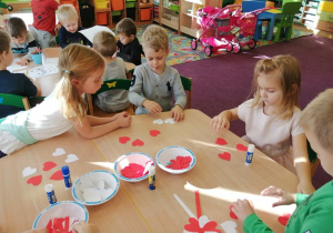 Grupa dzieci przy stolikach wykonuje z papierowych serduszek biało - czerwone kotyliony.